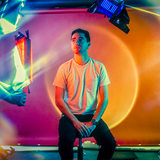 Colorful photograph of Kyle Finnegan, surrounded by film equipment, with the image tinted in neon colors of pink, yellow, green and blue.